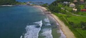 Vista aérea de uma praia com ondas, vegetação e casas próximas à costa.